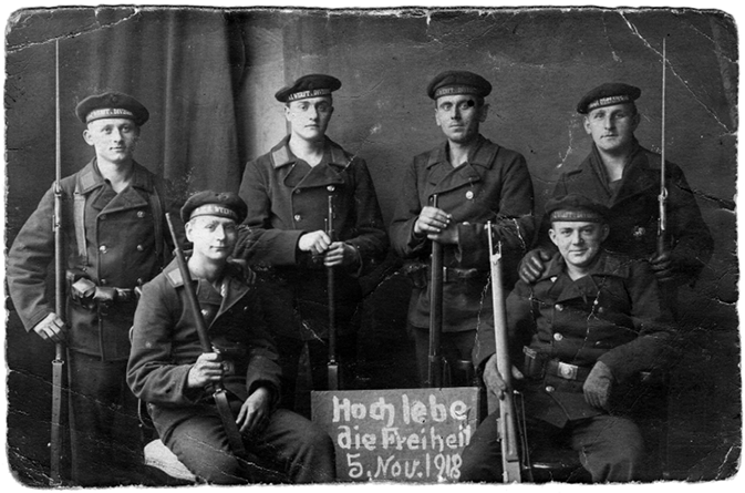 Six sailors armed with guns. They are grouped around a blackboard, which reads “Long live freedom 5. Nov. 1918”